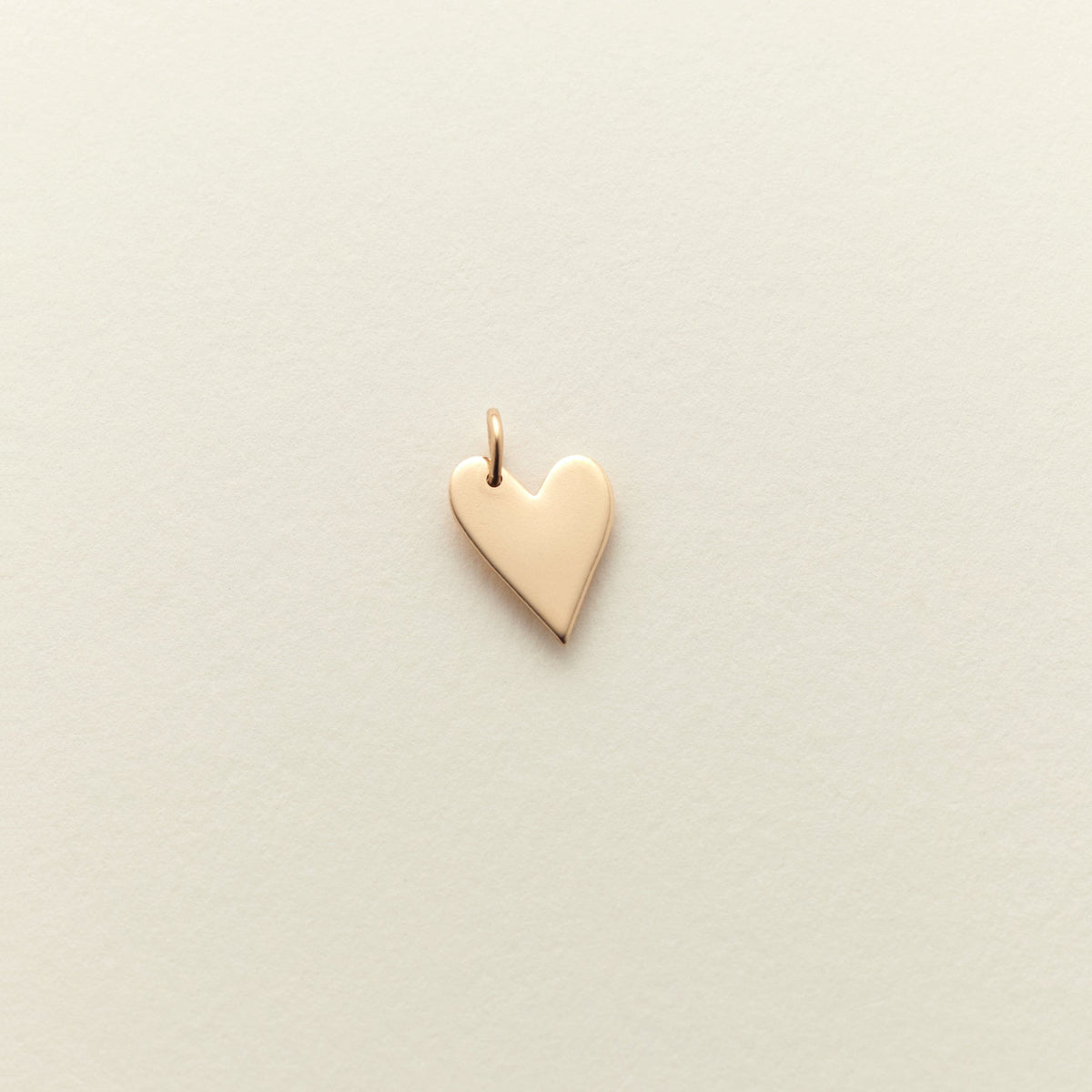A small, polished gold heart charm with a delicate top loop, centered on a textured off-white background.
