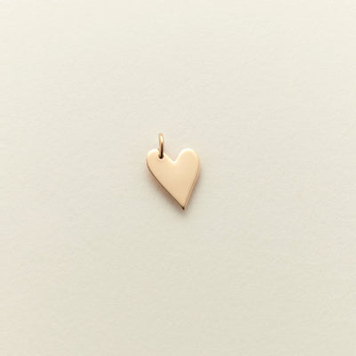 A small, polished gold heart charm with a delicate top loop, centered on a textured off-white background.