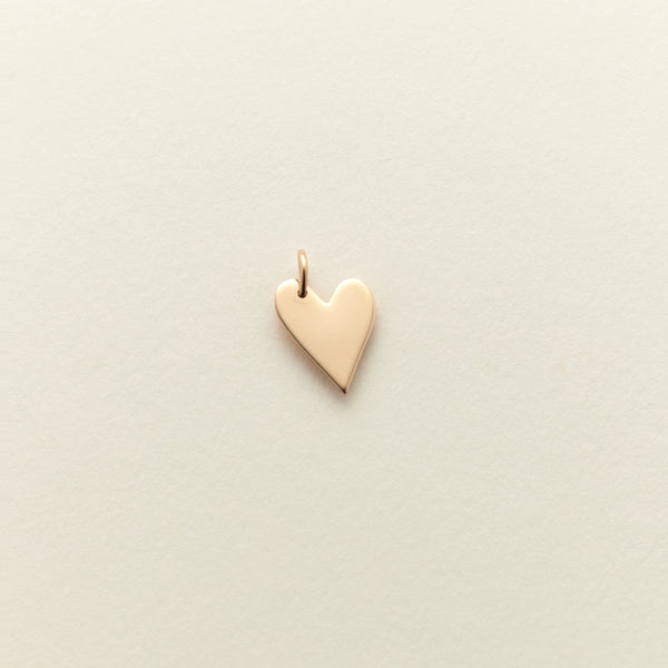 A small, polished gold heart charm with a delicate top loop, centered on a textured off-white background.