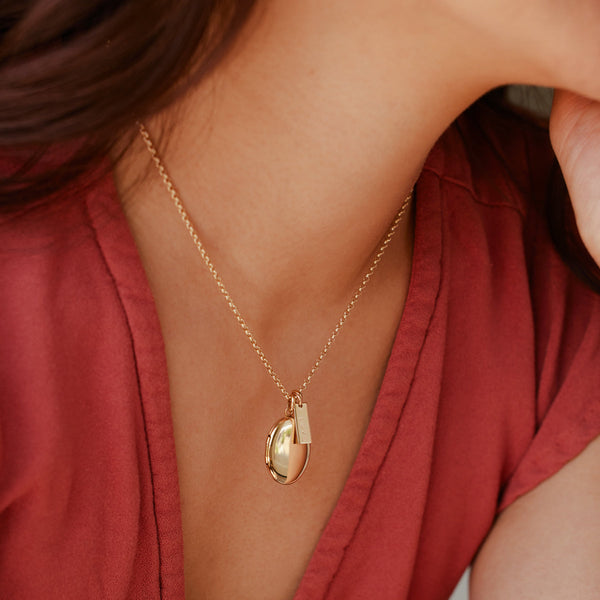 A close-up of a person wearing a red v-neck top paired with a gold chain necklace holding an oval locket charm and a small rectangular bar charm.