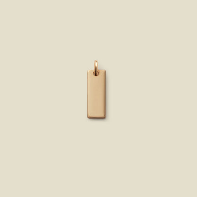 A small rectangular gold bar charm with a jump ring at the top, shown vertically on a solid beige background.