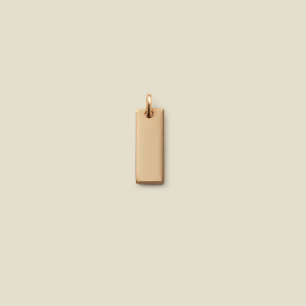 A small rectangular gold bar charm with a jump ring at the top, shown vertically on a solid beige background.