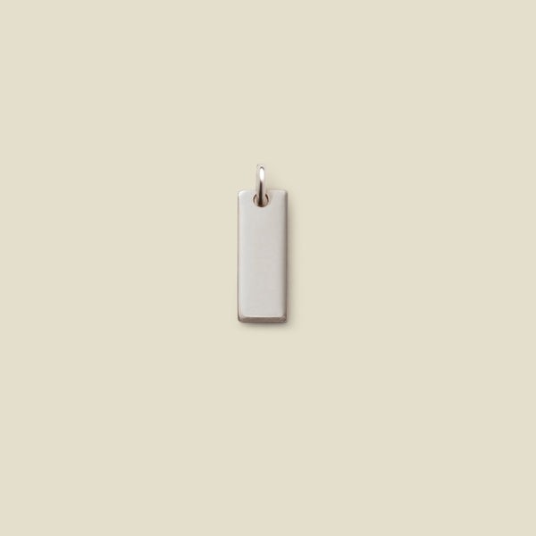A small rectangular silver bar charm with a jump ring at the top, shown vertically on a solid beige background.