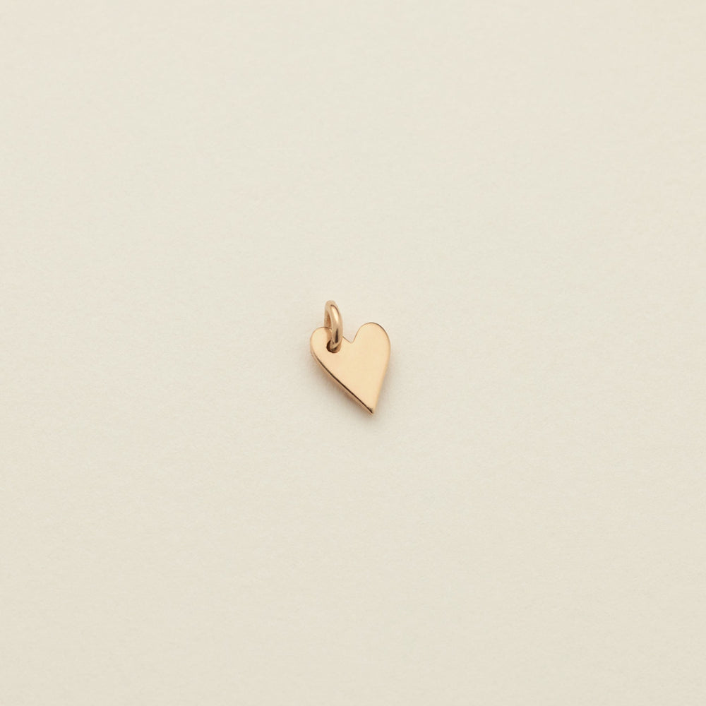 A small polished gold heart charm with a top loop, centered on a warm off-white textured background.