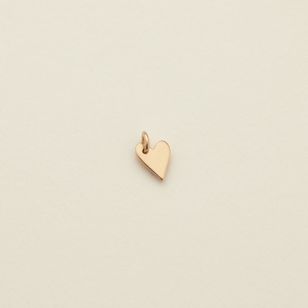 A small polished gold heart charm with a top loop, centered on a warm off-white textured background.