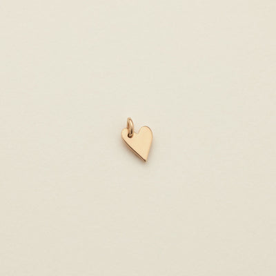 A small polished gold heart charm with a top loop, centered on a warm off-white textured background.