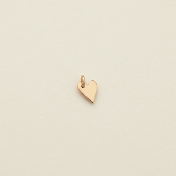 A small polished gold heart charm with a top loop, centered on a warm off-white textured background.