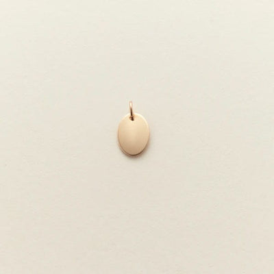 A small flat gold oval pendant, shown on a solid light beige background.