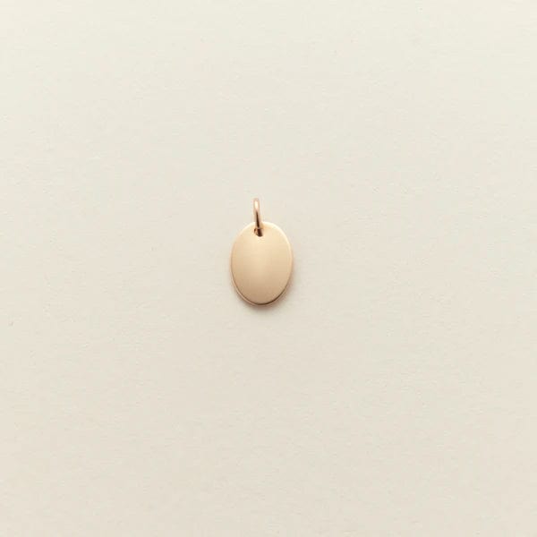 A small flat gold oval pendant, shown on a solid light beige background.
