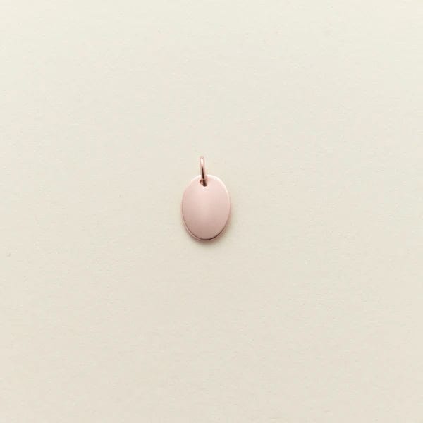A small flat rose gold oval charm, shown against a solid light beige background.