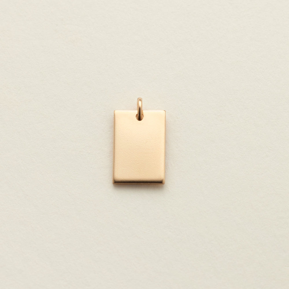 A small rectangular gold charm with a top loop, centered on a textured off-white background.