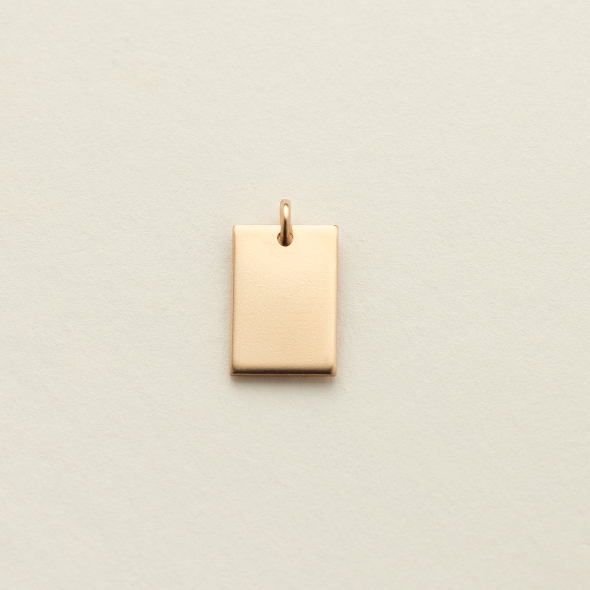 A small rectangular gold charm with a top loop, centered on a textured off-white background.