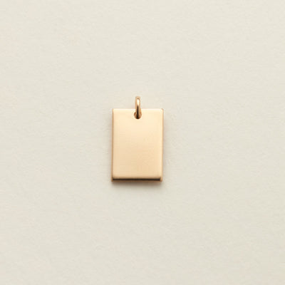 A small rectangular gold charm with a top loop, centered on a textured off-white background.
