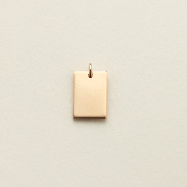 A small rectangular gold charm with a top loop, centered on a textured off-white background.