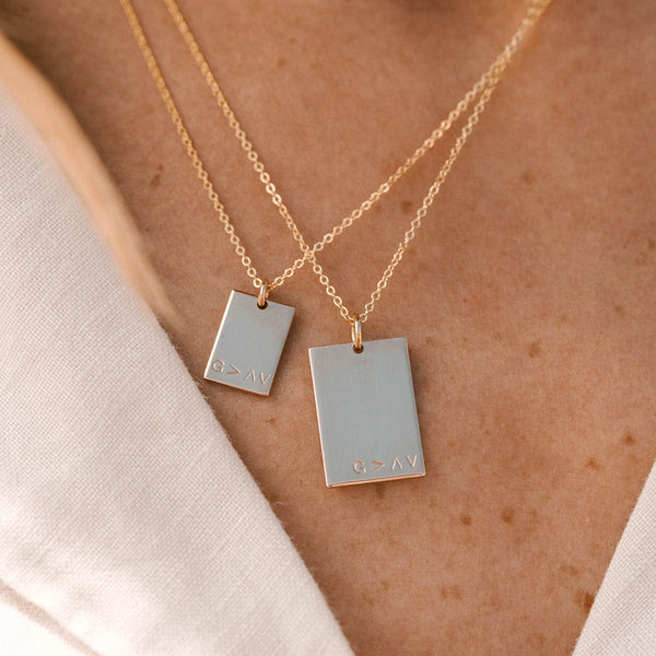 A close up of a person wearing two layered gold pendant chains, each with a different size of rectangle charm. Each charm is engraved with 'G>^v' along the bottom of the rectangle.