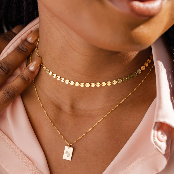 A close-up of a person wearing a gold choker with flat round discs linked together, and a delicate gold pendant chain with a floral engraved rectangle charm.