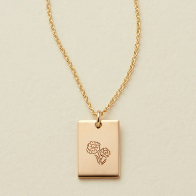 A delicate gold pendant chain necklace with a small polished rectangle charm, engraved with two flowers and shown on a textured light beige background.
