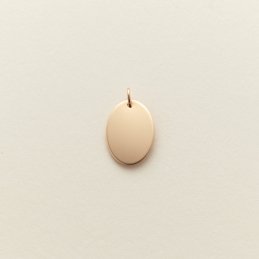 A small, blank oval pendant charm in a polished gold finish, centered on a textured off-white background.
