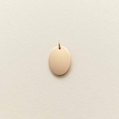 A small, blank oval pendant charm in a polished gold finish, centered on a textured off-white background.