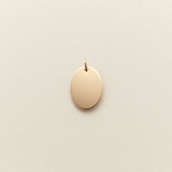 A small, blank oval pendant charm in a polished gold finish, centered on a textured off-white background.
