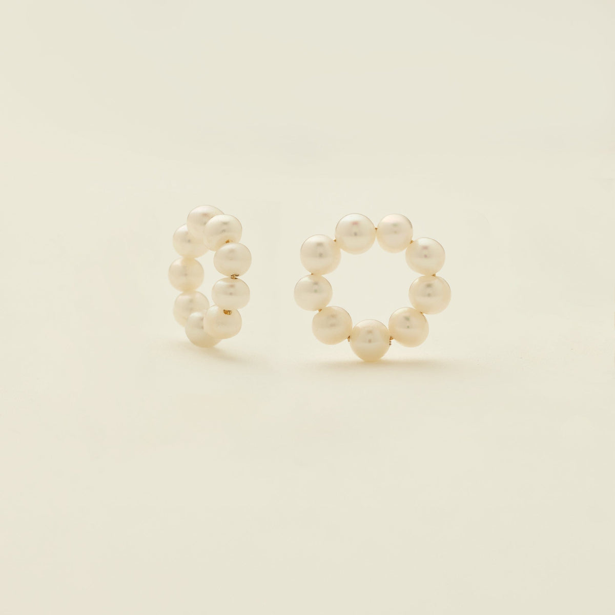 A pair of round ring hoop earring attachments, made-up of round cream colored pearls, shown on a solid light beige background.