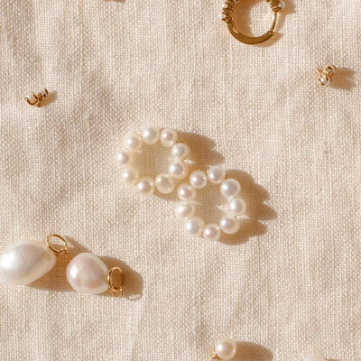 A pair of round ring hoop earring attachments, made-up of round cream colored pearls, laying centered on a cream colored linen surface with other pearl jewelry surrounding them.