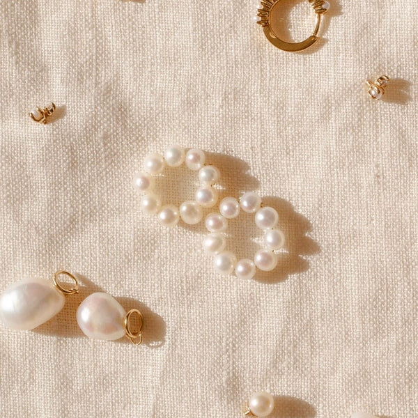 A pair of round ring hoop earring attachments, made-up of round cream colored pearls, laying centered on a cream colored linen surface with other pearl jewelry surrounding them.