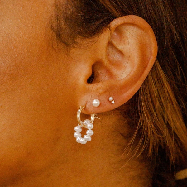 A close-up of a person's ear wearing a small gold hoop earring with a round ring of pearls dangling from it, a simple pearl stud, and a gold stud earring with a small pearl and a small clear accent stone.