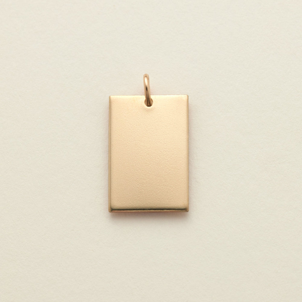 A polished gold rectangle pendant with a top loop, shown on a solid light beige background.