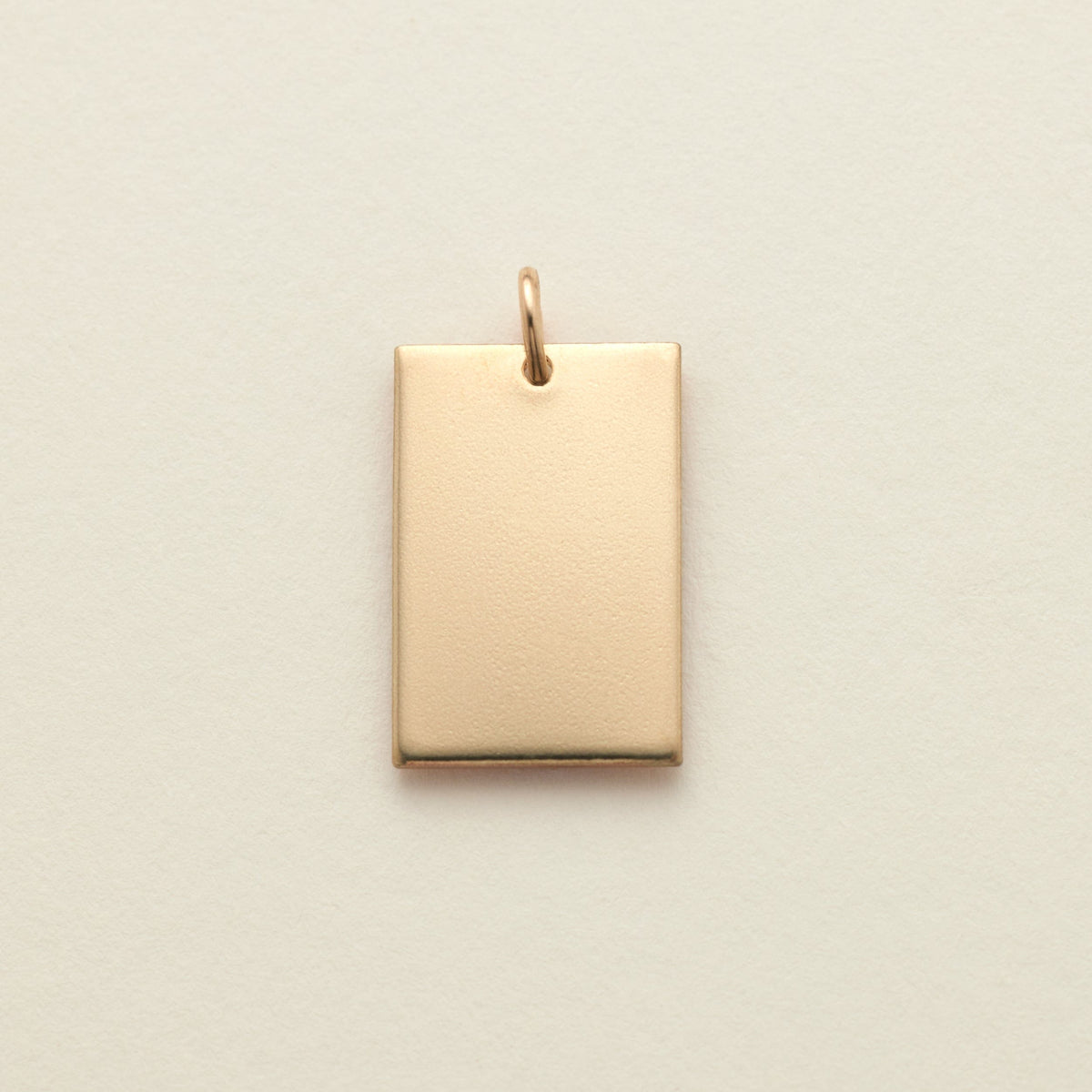 A polished gold rectangle pendant with a top loop, shown on a solid light beige background.