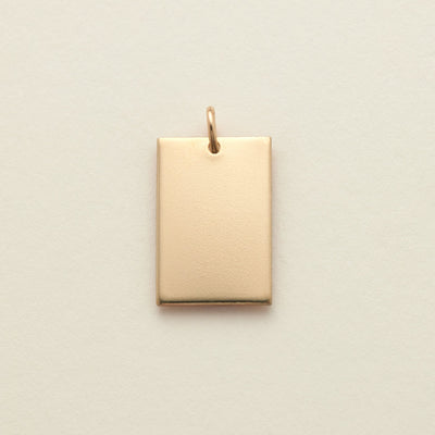 A polished gold rectangle pendant with a top loop, shown on a solid light beige background.