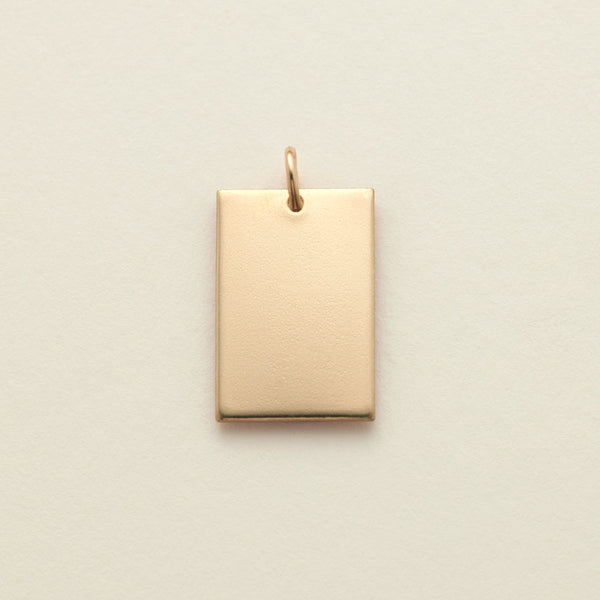 A polished gold rectangle pendant with a top loop, shown on a solid light beige background.