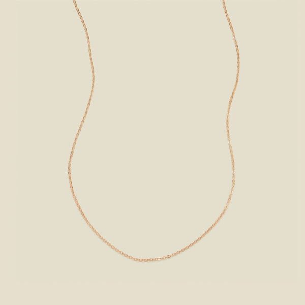 A delicate rose gold chain necklace arranged in a U-shape against a solid, warm beige background.