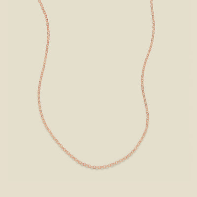 A delicate rose gold chain necklace arranged in a U-shape against a solid, warm beige background.