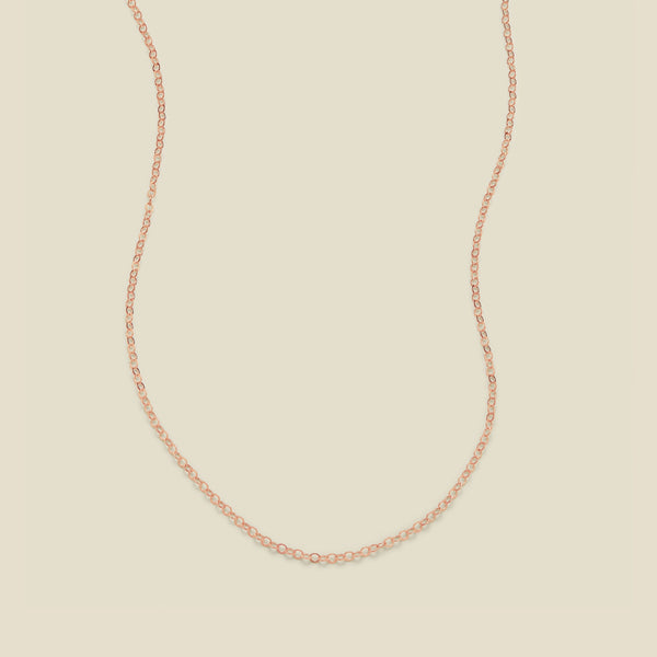 A delicate rose gold chain necklace arranged in a U-shape against a solid, warm beige background.
