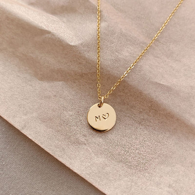 A delicate gold pendant chain with a round flat disc charm, engraved with the letter 'M' and a heart, laying on a textured light beige surface.