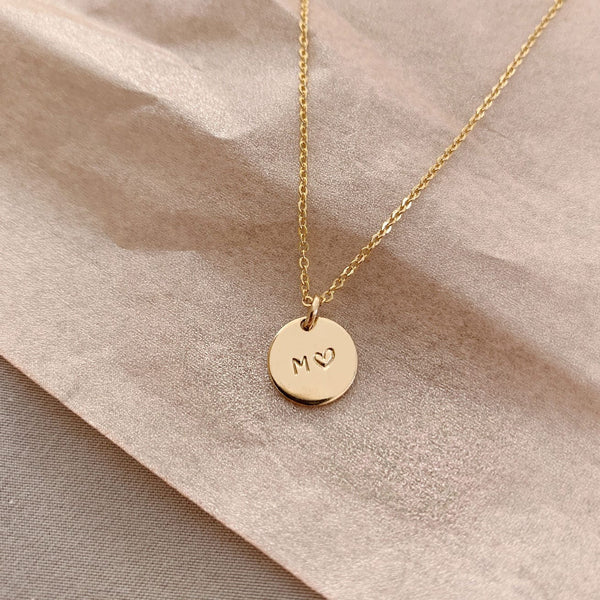A delicate gold pendant chain with a round flat disc charm, engraved with the letter 'M' and a heart, laying on a textured light beige surface.