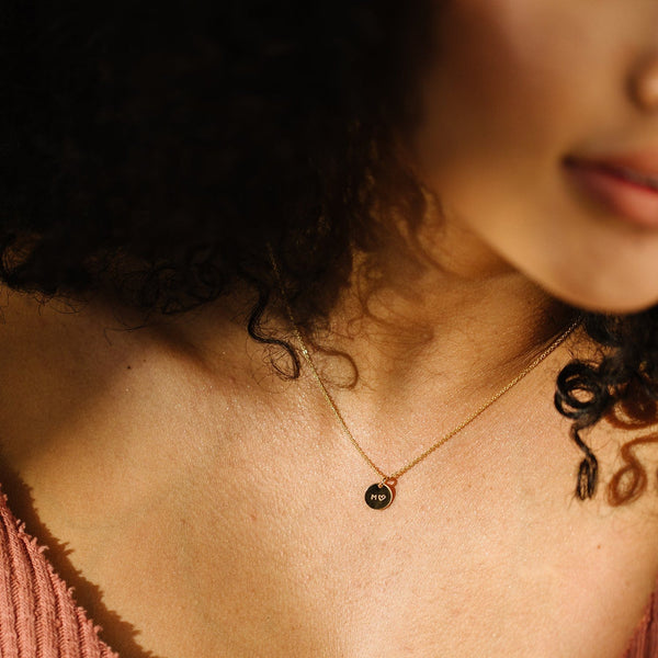 A close-up of a person's neck wearing a delicate gold pendant chain with a round engraved flat disc charm.