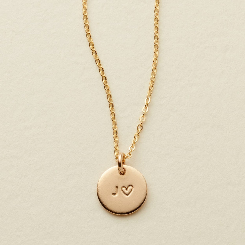 A delicate gold chain necklace with a round gold disc pendant engraved with the initials 'J' and a heart, displayed on a textured off-white background.