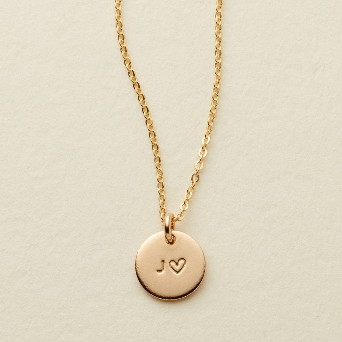 A delicate gold chain necklace with a round gold disc pendant engraved with the initials 'J' and a heart, displayed on a textured off-white background.