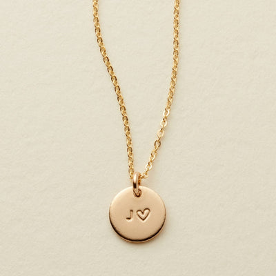 A delicate gold chain necklace with a round gold disc pendant engraved with the initials 'J' and a heart, displayed on a textured off-white background.