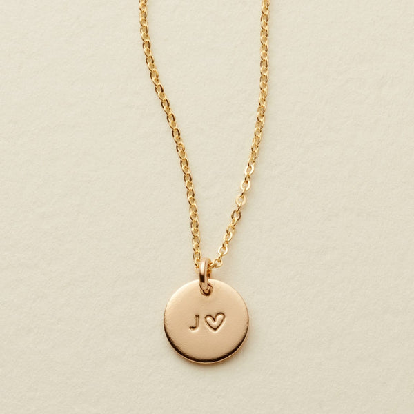 A delicate gold chain necklace with a round gold disc pendant engraved with the initials 'J' and a heart, displayed on a textured off-white background.