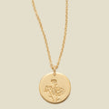 A gold chain necklace with a round gold disc pendant featuring an engraved daisy flower design, displayed against a solid light beige background.