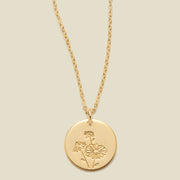 A gold chain necklace with a round gold disc pendant featuring an engraved daisy flower design, displayed against a solid light beige background.