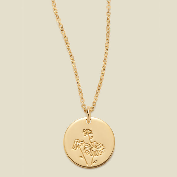 A gold chain necklace with a round gold disc pendant featuring an engraved daisy flower design, displayed against a solid light beige background.