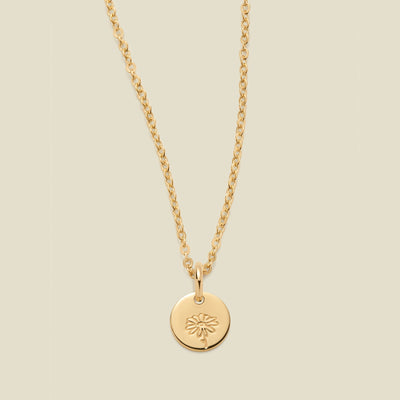 A gold chain necklace with a round gold disc pendant featuring an engraved daisy flower design, displayed against a solid light beige background.