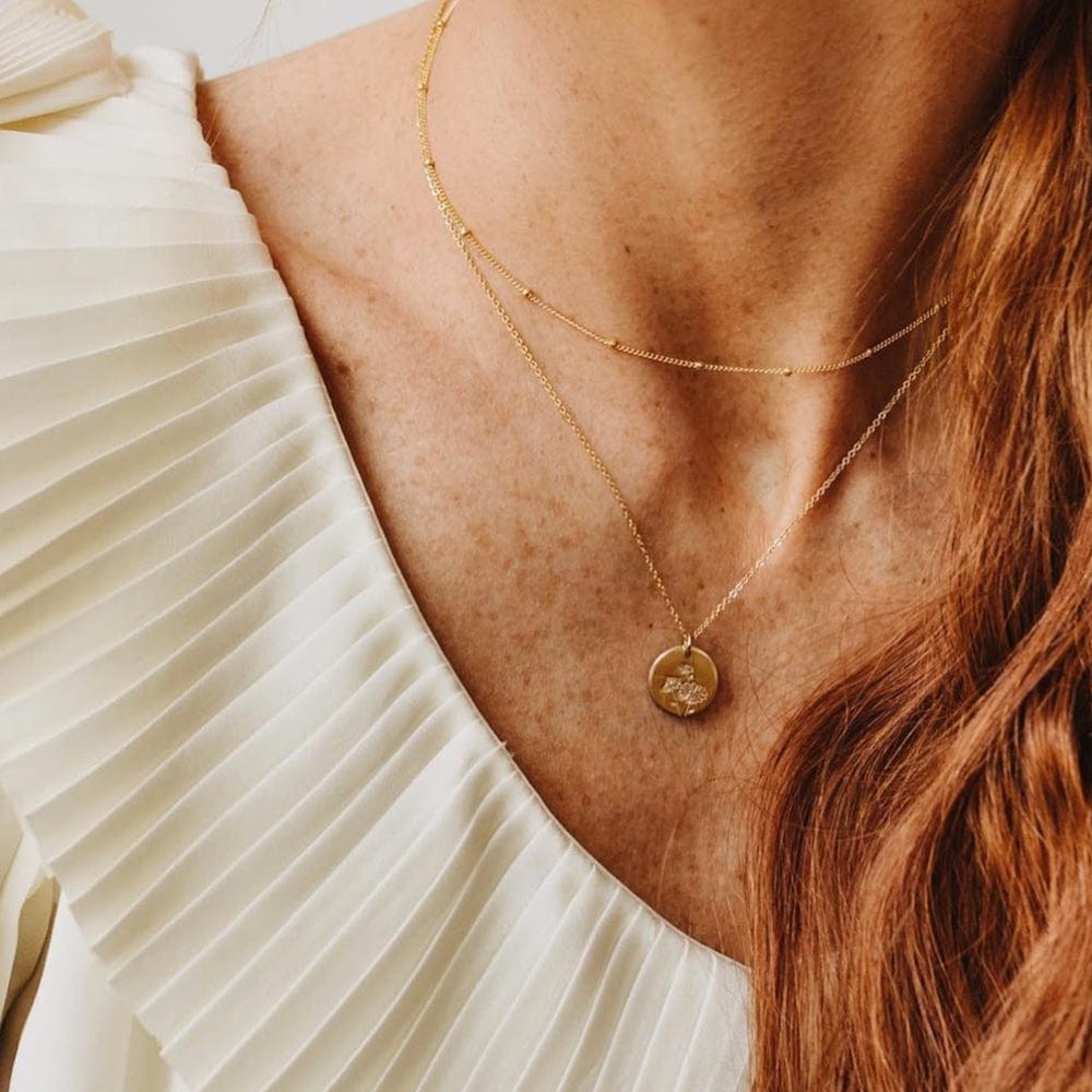 A close-up of a woman in a white pleated top wearing a thin gold necklace with stationary round beads layered with a delicate pendant chain featuring a round flat disc charm. The charm is engraved with three small daisy flowers.