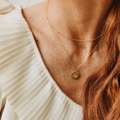 A close-up of a woman in a white pleated top wearing a thin gold necklace with stationary round beads layered with a delicate pendant chain featuring a round flat disc charm. The charm is engraved with three small daisy flowers.