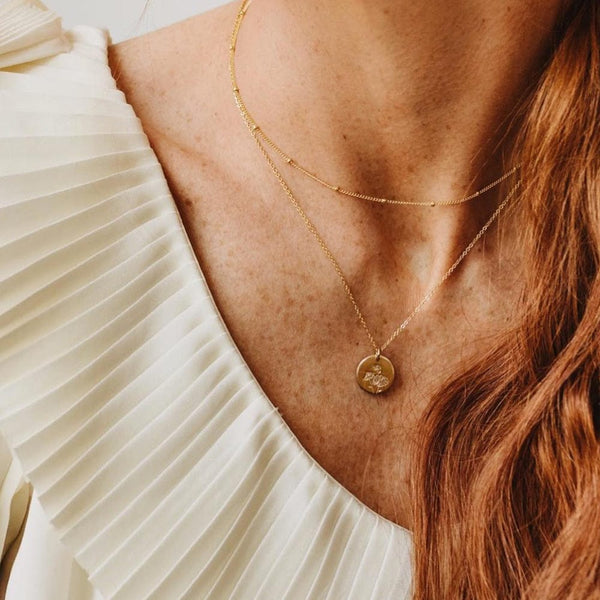 A close-up of a woman in a white pleated top wearing a thin gold necklace with stationary round beads layered with a delicate pendant chain featuring a round flat disc charm. The charm is engraved with three small daisy flowers.