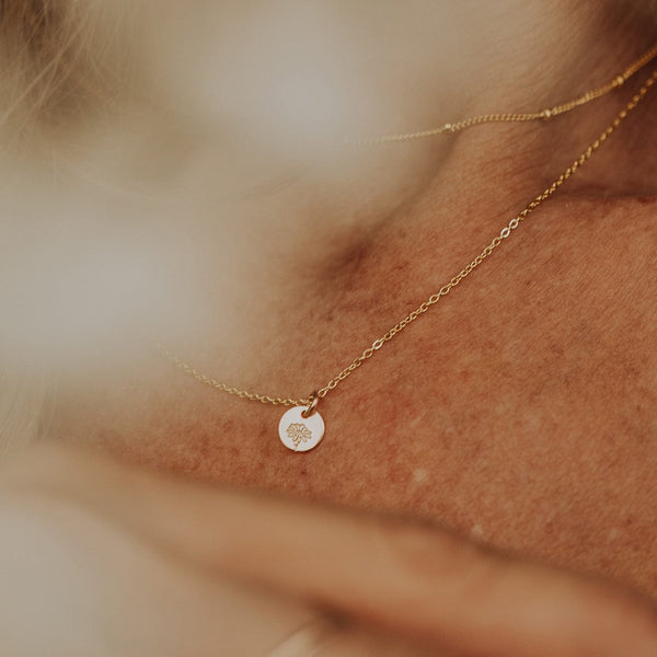 A close-up of a person wearing a delicate gold chain necklace with a flat round disc, engraved with a daisy flower design.